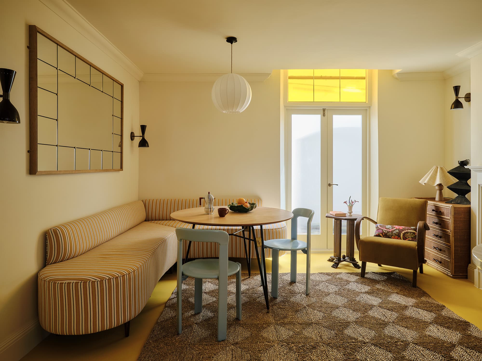 A retro-style dining and seating area in a warm yellow room, featuring a curved striped banquette, a round wooden table with light blue chairs, a patterned rug, and a yellow armchair near a window.