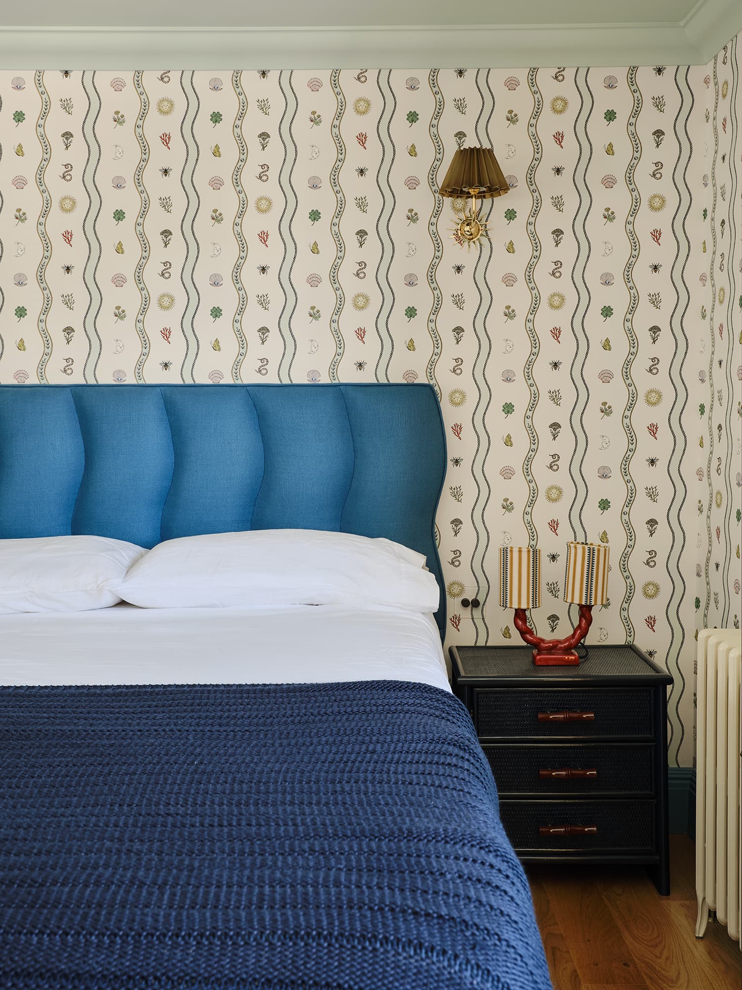 A styled bedroom corner featuring a bed with a prominent blue tufted headboard, white bedding, a navy blue textured blanket, a black nightstand, and a wall covered in cream wallpaper with vertical wavy lines and small multicolored illustrations.