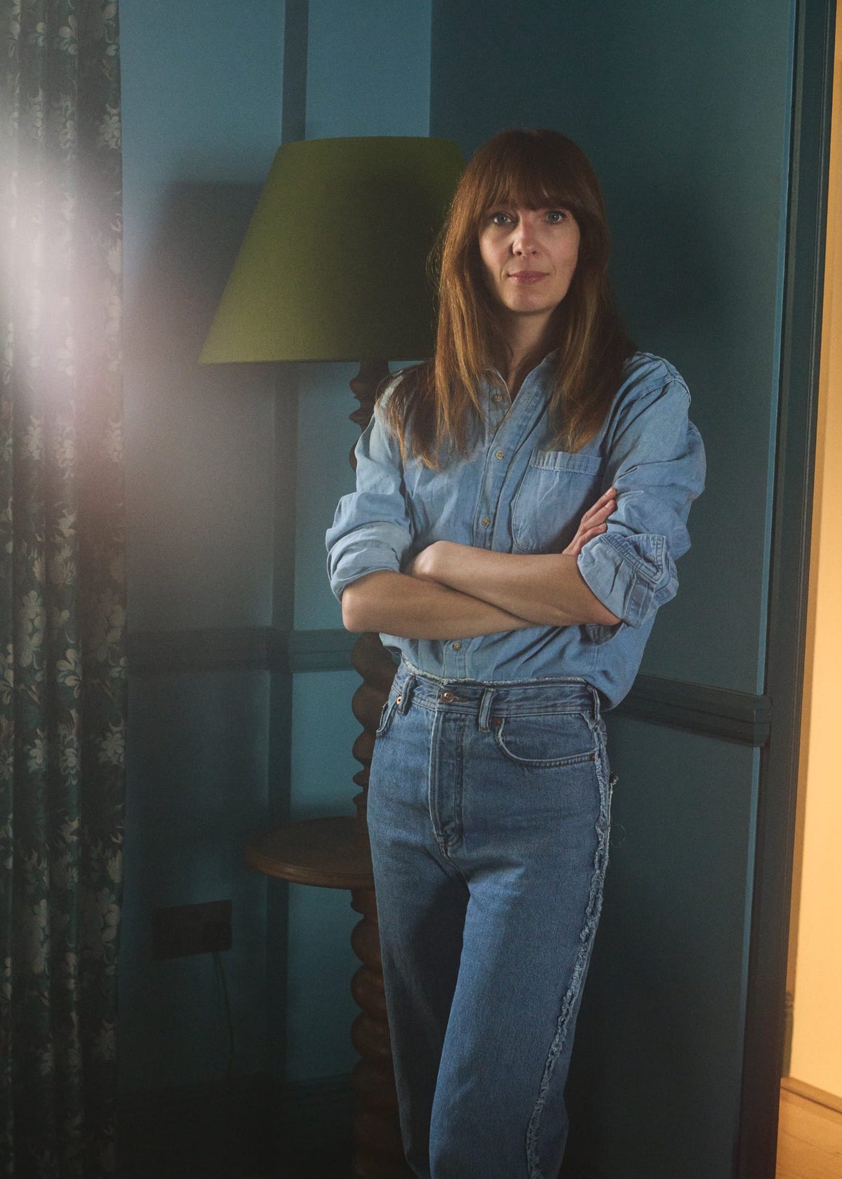 A woman with shoulder-length brown hair stands indoors with her arms crossed, wearing a light blue denim button-up shirt and matching blue jeans. She is posed in a room with dark teal walls, a green lamp, and a curtain on the left.