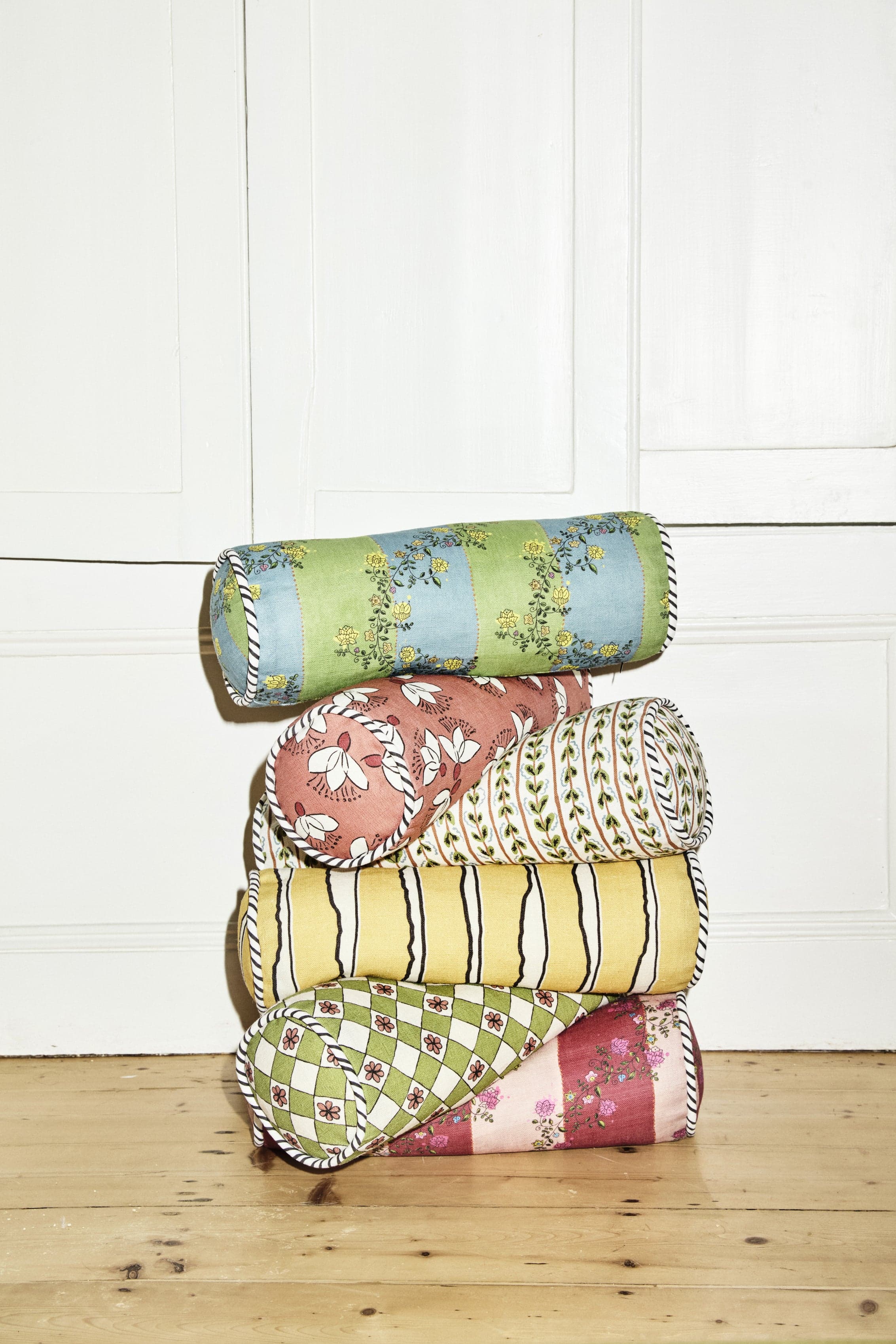 A stack of four rolled fabric cushions or bolsters in different floral and striped patterns, set on a light wooden floor against a white paneled wall.