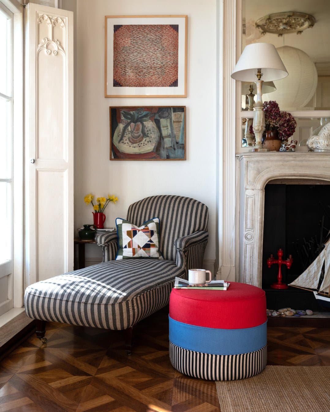 A styled interior corner featuring a striped chaise lounge with a patterned pillow, a colorful striped ottoman, framed wall art, a fireplace, and wood parquet flooring.